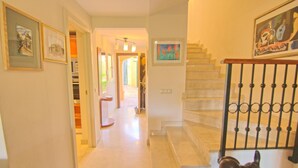 A hallway with a staircase leading to a landing. The hallway has framed pictures on the walls and a door at the end.