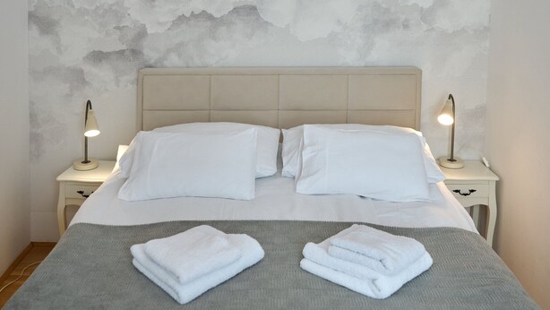 A neatly made bed with white linens and two folded towels, a textured gray wall, and a modern pendant light fixture. - Codelli Deluxe Unique Fresko Apartments (Ljubljana)