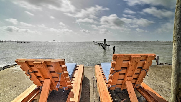 Twee houten lounge fauteuils met blauwe kussens op een betonnen terras aan het water, met een steiger die zich uitstrekt in de zee.