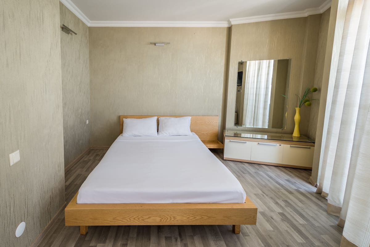 A neatly made bed with a wooden frame and white bedding, a bedside table with a mirror, and a small vase with green plants.