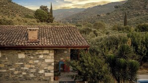 Un bâtiment en pierre, surmonté d’un toit en tuiles, entouré de végétation et de collines.