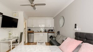 A compact bedroom with a bed, a grey sofa, a television, a ceiling fan, a washing machine, and a kitchenette with white cabinets and a microwave.