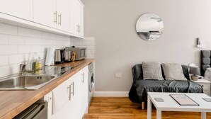 A compact kitchen with white cabinets, a sink, and a microwave. There's a round mirror on the wall and a bed with pillows in the background.