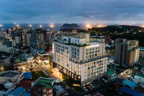 A multi-story hotel with a rooftop terrace, surrounded by other buildings and a hilly landscape in the background. - Shin Shin Hotel Cheonjiyeon (Seogwipo)