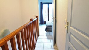 A narrow hallway with a wooden railing, a door on the right, and a view into a room with a bed and curtains.