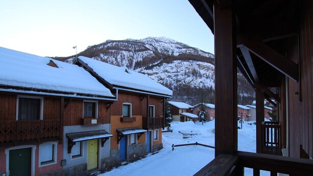 A snow-covered street with wooden buildings, a mountain in the background, and a wooden balcony railing.