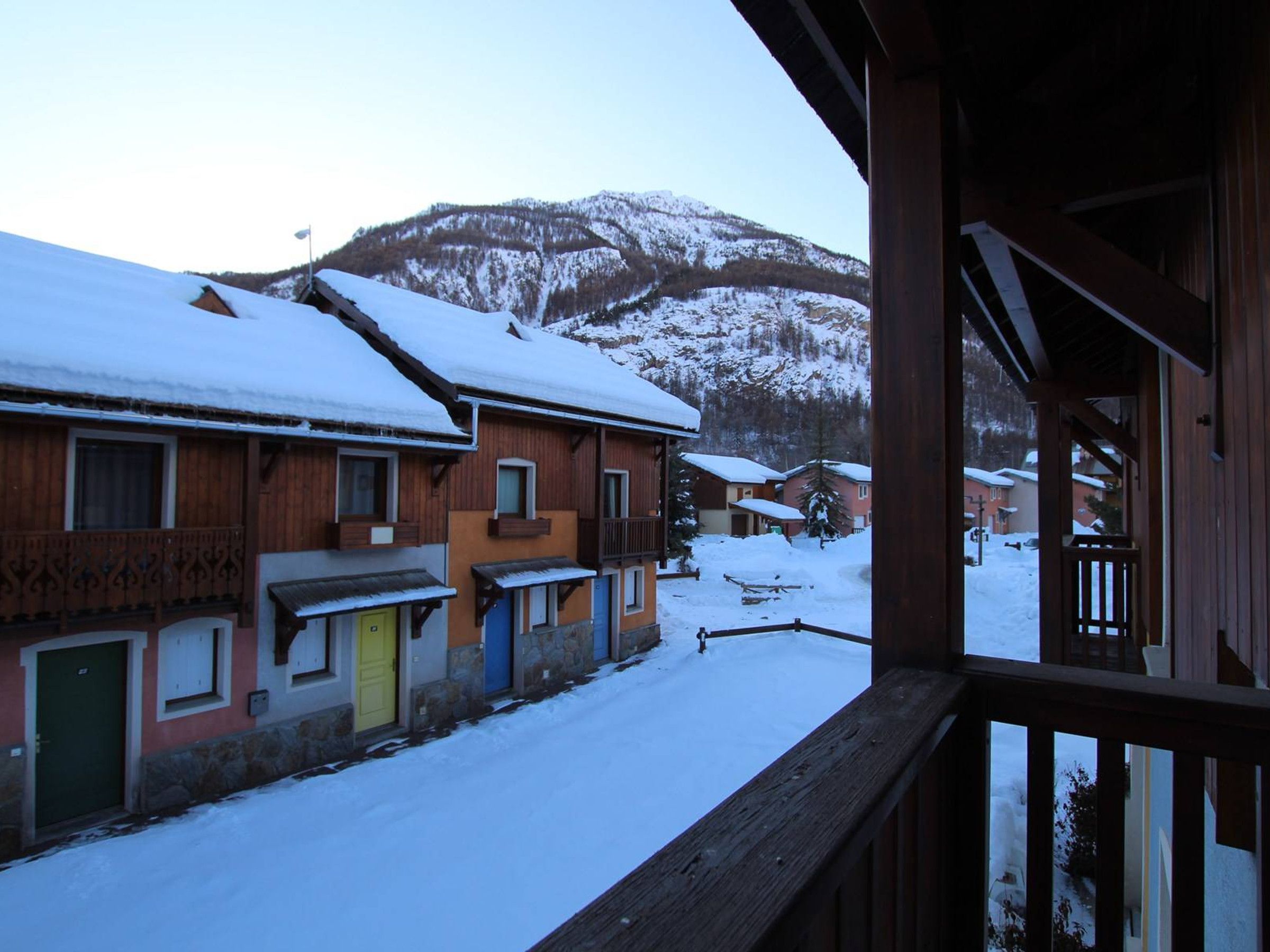 Eine verschneite Straße mit Holzbauten, ein Berg im Hintergrund und ein hölzernes Balkongeländer.