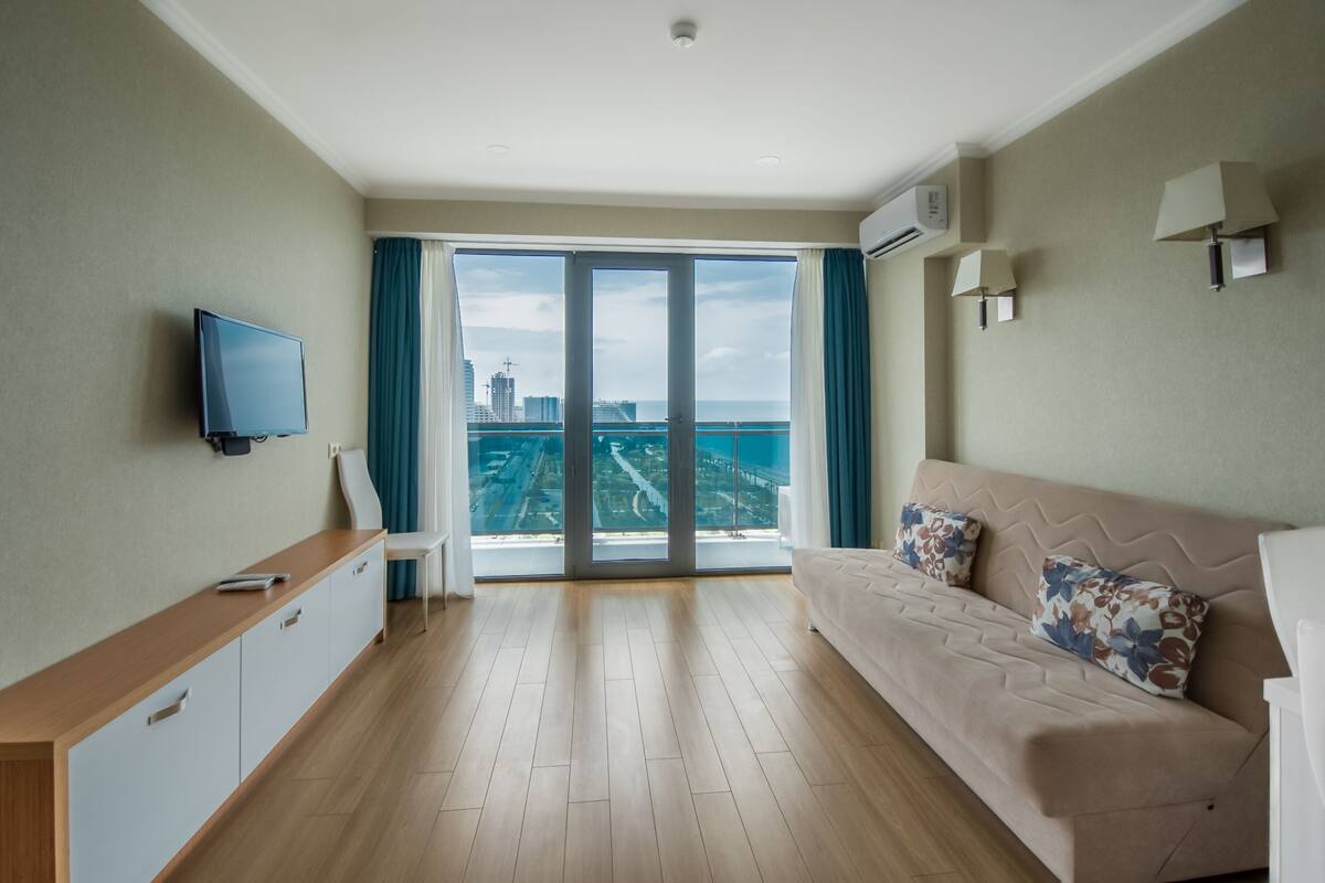Un salon moderne comprenant un canapé, un téléviseur, un bureau et une grande porte-fenêtre coulissante donnant sur un balcon avec vue sur la ville.
