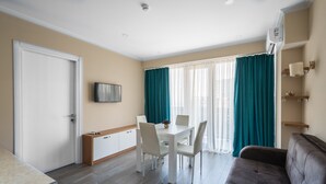 A modern living room with a sofa, a dining table with chairs, a TV mounted on the wall, and a window with curtains. - Orbi Beach Tower Hotel Official (Batumi)