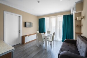 Una sala de estar moderna con un sofá, una mesa de comedor con sillas, un televisor montado en la pared y un ventanal con cortinas.