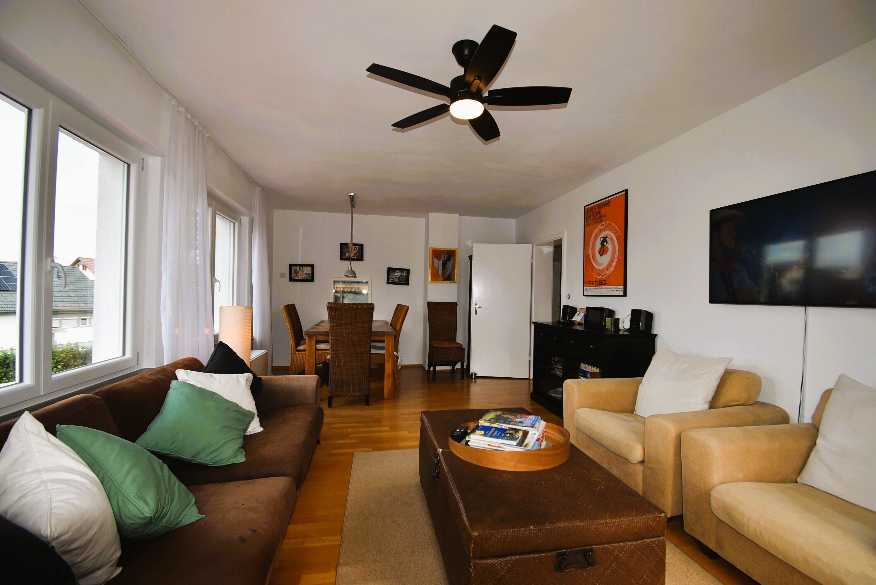 Una sala de estar con un sofá marrón, un sofá beige y una mesa de centro con libros. Hay un ventilador de techo, una mesa de comedor con sillas y un televisor montado en la pared.