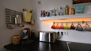 A kitchen with a toaster, coffee maker, and various kitchen utensils. There are shelves with bottles and a hanging rack with cups. - Sunny 4 room apartment near the lake with south-facing roof terrace
 (Eriskirch)
