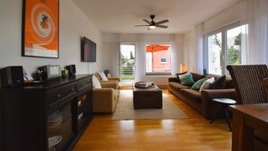 A living room with a sofa, ottoman, and a television on a cabinet. There is a ceiling fan and a window with curtains. - Sunny 4 room apartment near the lake with south-facing roof terrace
 (Eriskirch)