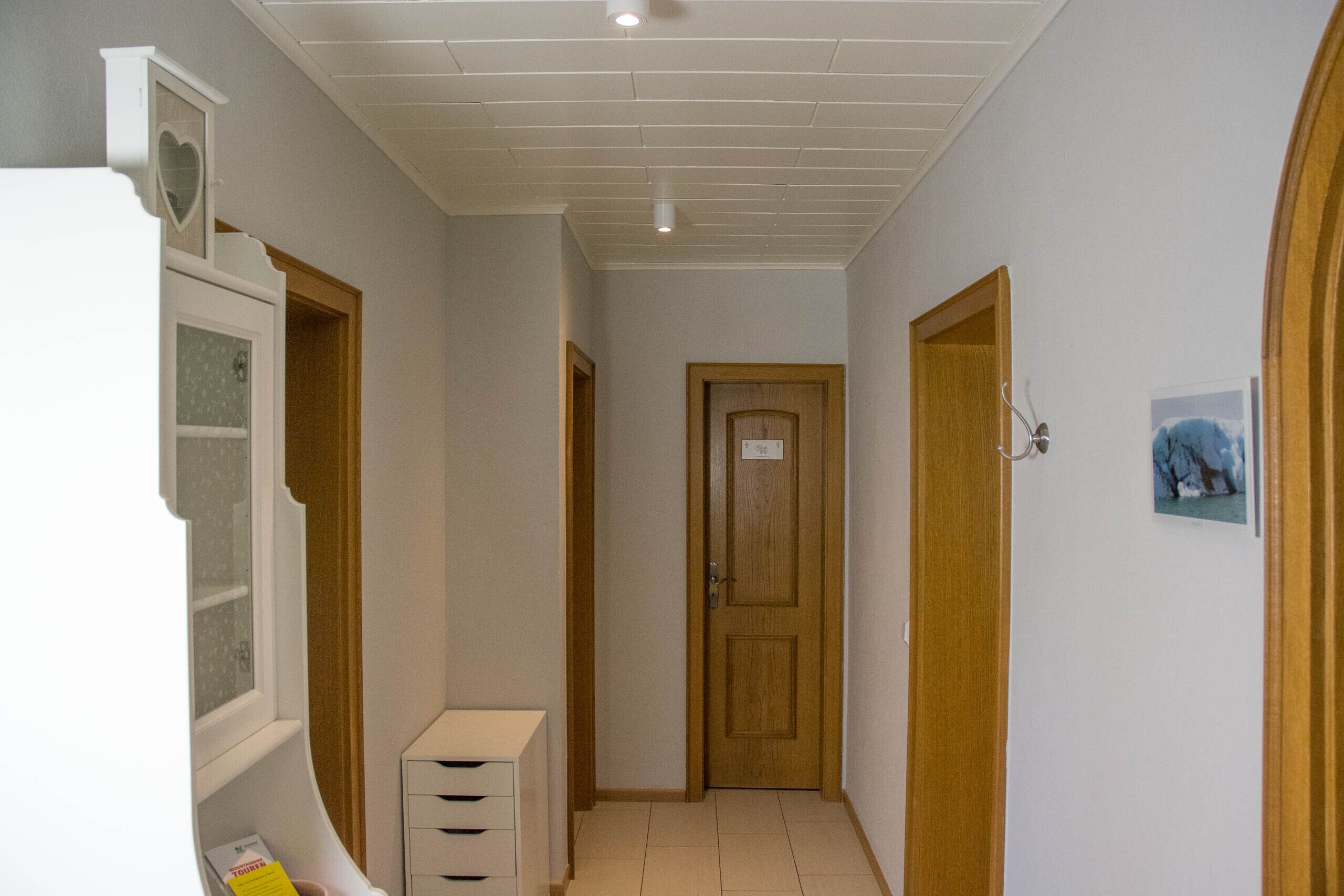 A narrow hallway with wooden doors, a white wall, and a ceiling with recessed lighting.