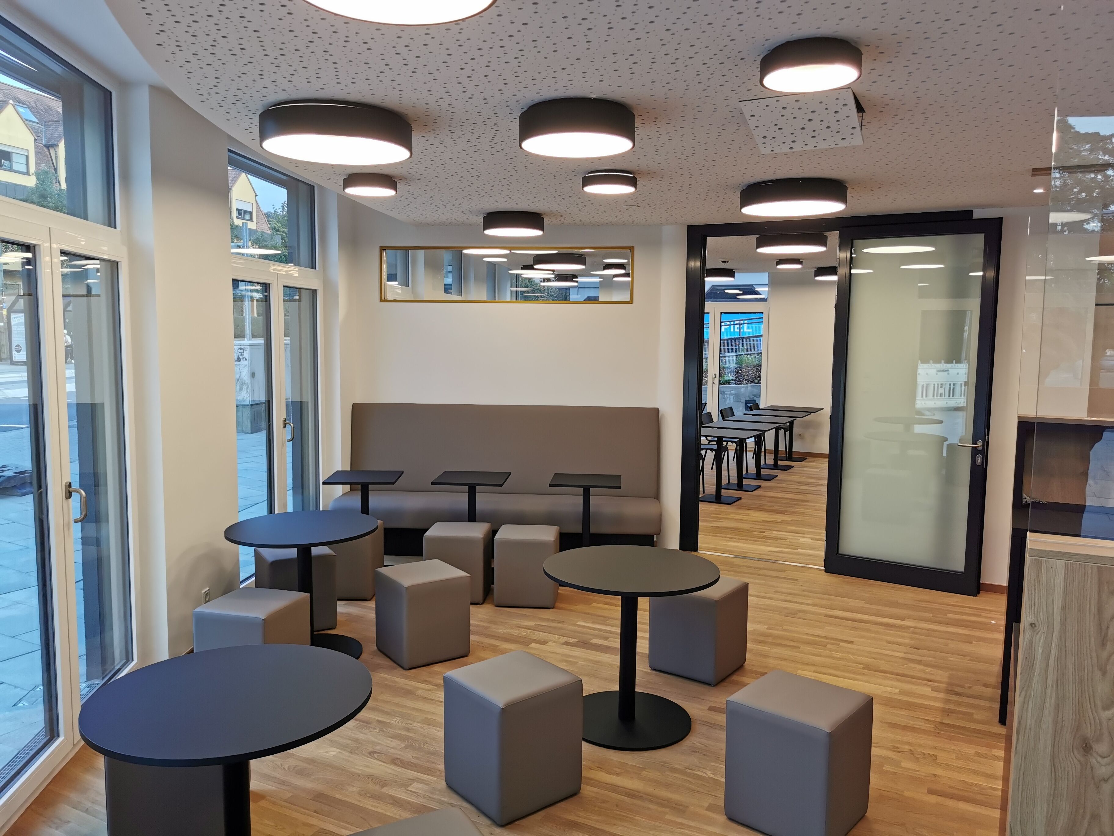 A modern interior with round tables and grey seating, large windows, and a wooden floor.