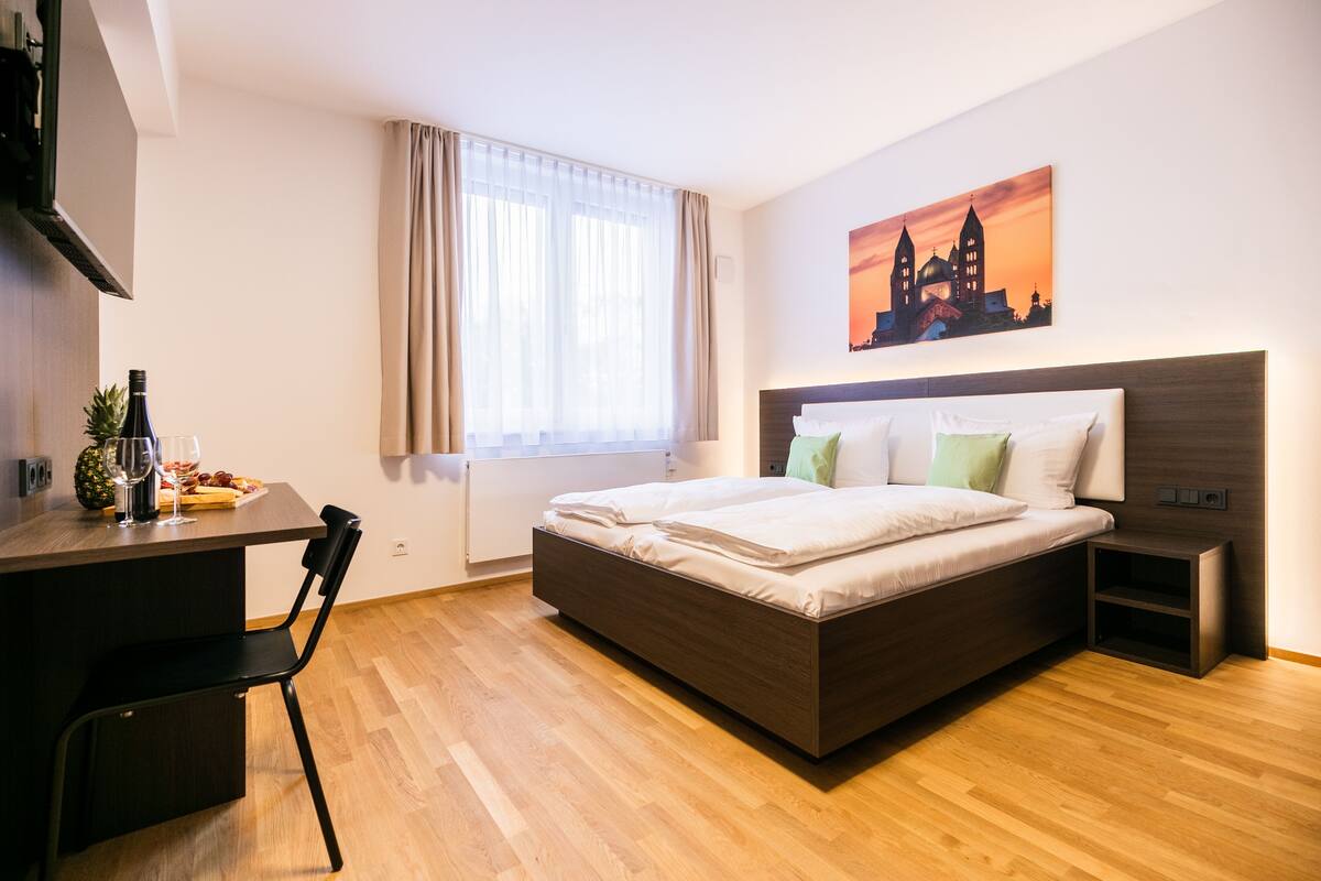A modern hotel room with a wooden floor, a bed with white linens, a desk with a chair, a small table with a bottle and fruit, and a painting of a cityscape on the wall.