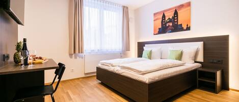 A modern hotel room with a wooden floor, a bed with white linens, a desk with a chair, a small table with a bottle and fruit, and a painting of a cityscape on the wall.