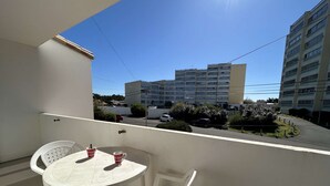 A balcony with a white table and two chairs, overlooking a street with parked cars and buildings. - Cozy 1st-Floor Apartment, 200m from Beach, Balcony, Parking & WiFi Options - Saint-Hilaire-de-Riez (Saint-Hilaire-de-Riez)