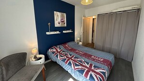 A bedroom with a bed, a chair, a bedside table, a lamp, a hanging light fixture, a door, and a curtain. - Cozy 1st-Floor Apartment, 200m from Beach, Balcony, Parking & WiFi Options - Saint-Hilaire-de-Riez (Saint-Hilaire-de-Riez)