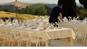 Mesa de comedor al aire libre con manteles y sillas blancos, un camarero al fondo y una vista panorámica de colinas y árboles.