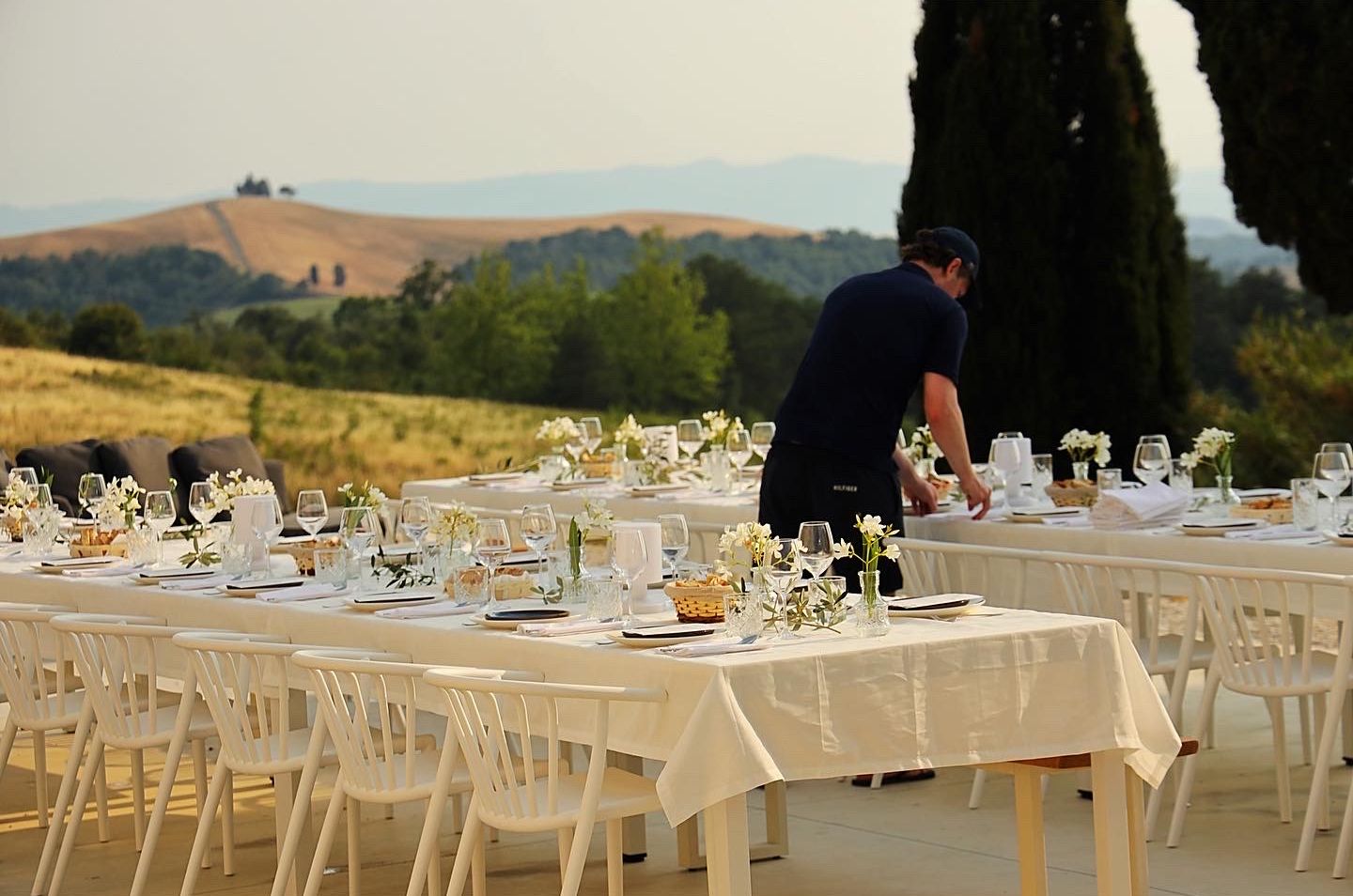 An outdoor dining setup with white tablecloths and chairs, a waiter in the background, and a scenic view of hills and trees.