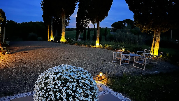 Un sendero de grava que conduce a un área de descanso con sillas y una mesa, rodeado de árboles y un arreglo floral.
