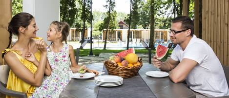 A family dining outdoors at a round table with a fruit basket, plates, and a view of a building and trees.