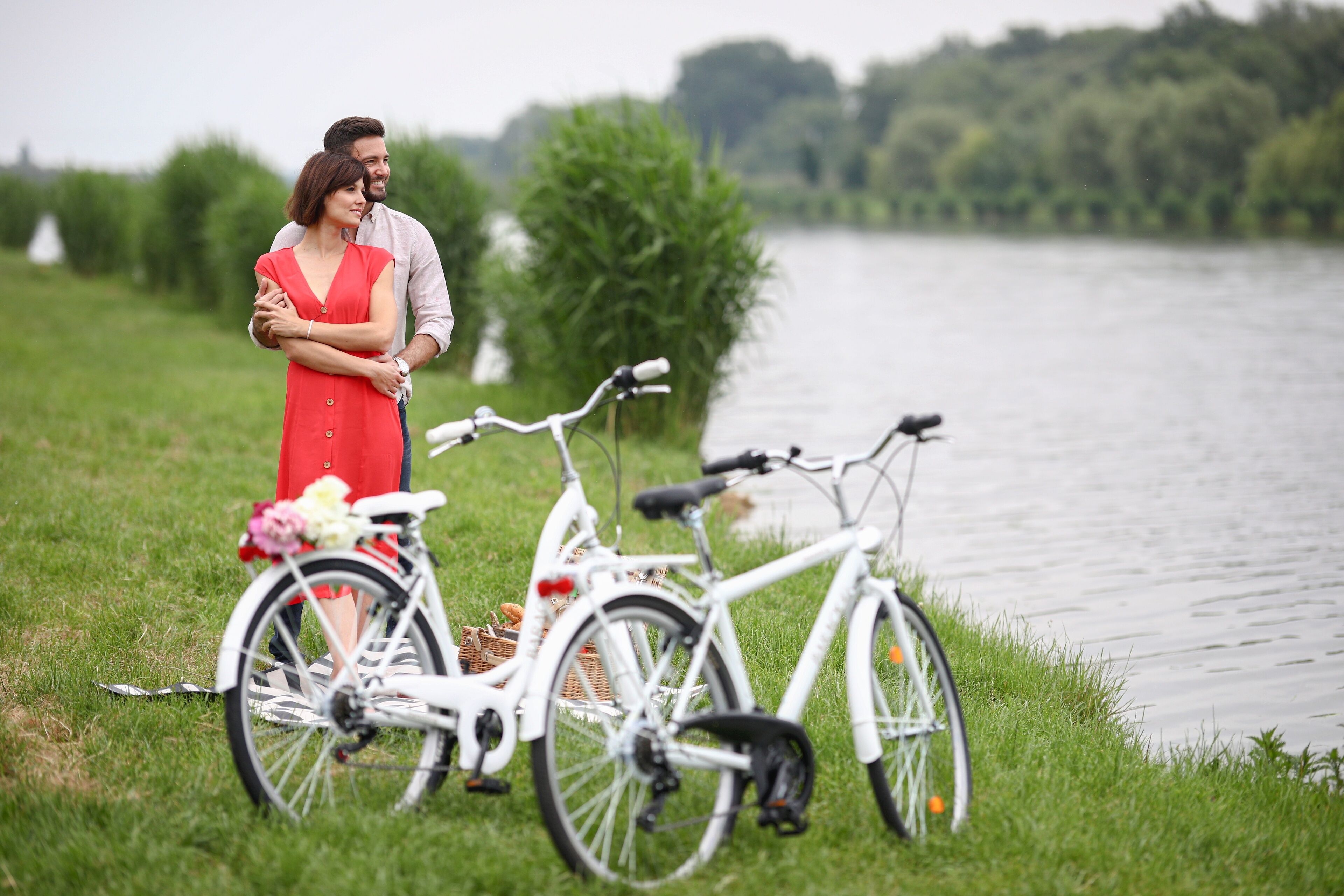 Una pareja de pie junto a una bicicleta blanca adornada con flores, cerca de agua y follaje.