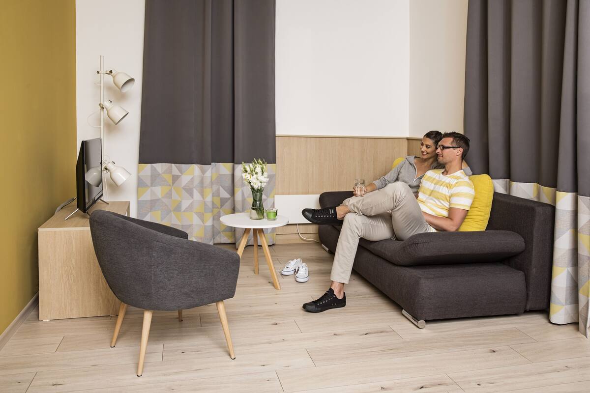 Ein modernes Wohnzimmer mit einer Couch, einem Sessel, einem kleinen runden Tisch mit Blumenvase und einem Fernseher auf einem Schrank.