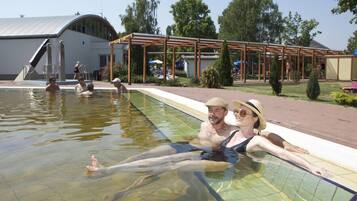 A swimming pool with people in the water, a covered area with umbrellas, and a building with a curved roof in the background.