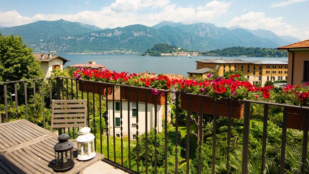 Un balcón con vista a un lago y montañas, una mesa de madera con un farol, y macetas con flores rojas.
