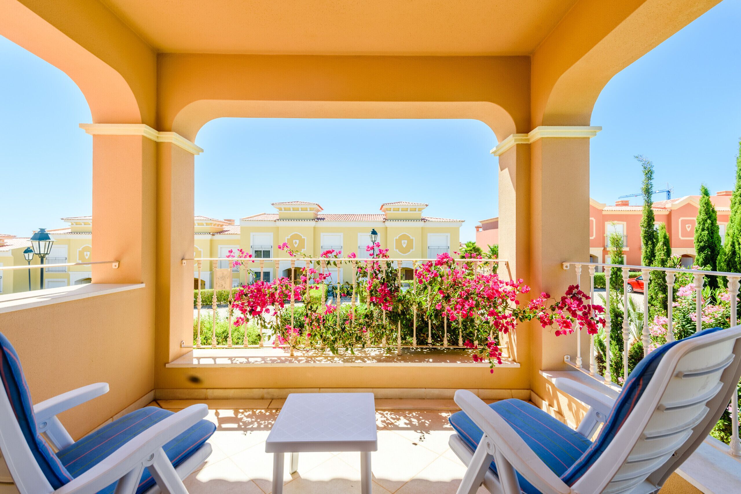 Un balcon avec deux fauteuils de jardin blancs, une petite table et une vue sur des bâtiments et des fleurs roses en arrière-plan.