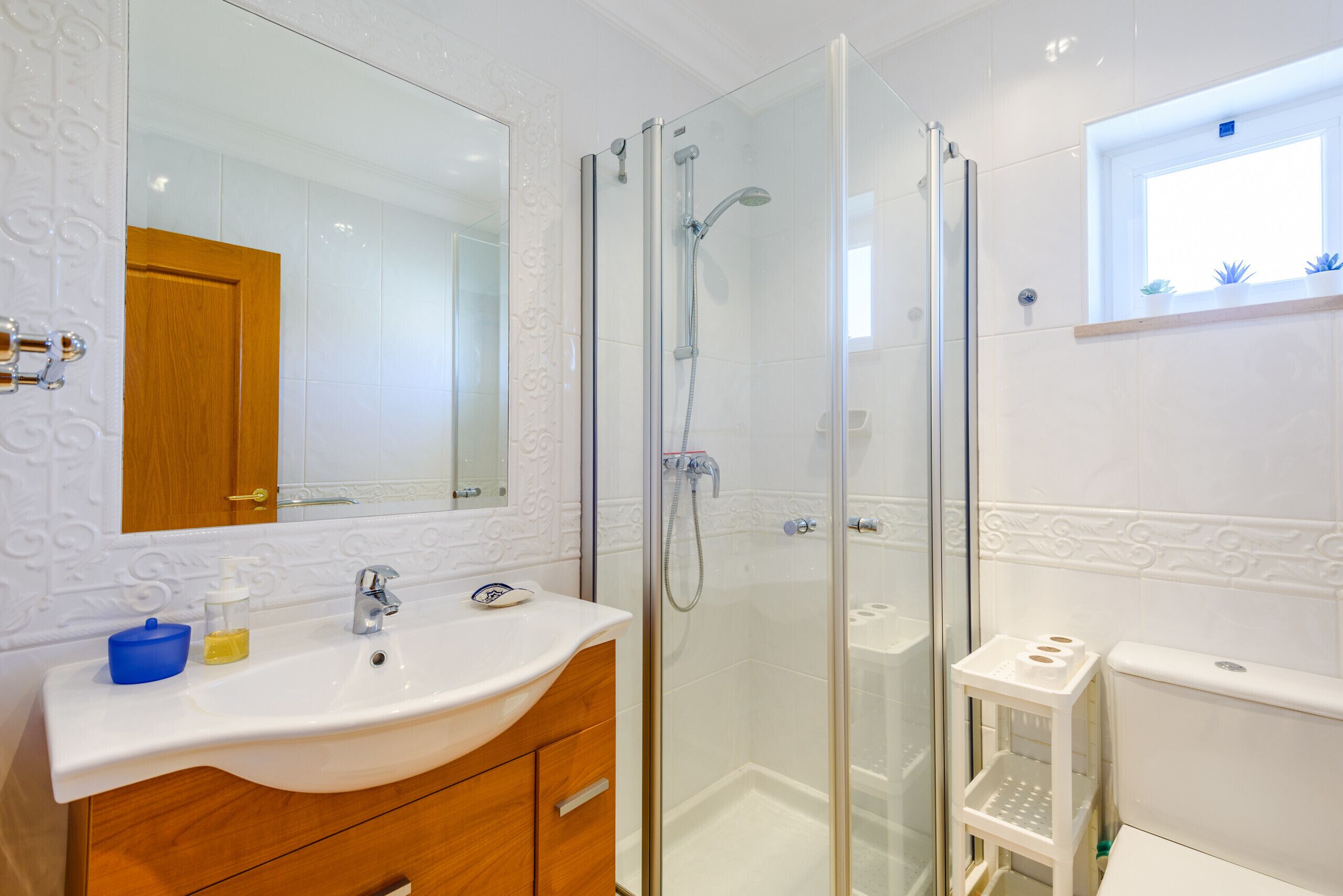 Un baño moderno con ducha de vidrio, lavamanos blanco sobre tocador de madera y un inodoro con un estante pequeño.
