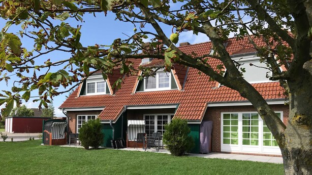 A two-story house with a red-tiled roof, a front porch with chairs, and a well-maintained lawn. - Langes Hus 2 - Ferienhäuser Langes Hus (Friedrichskoog)