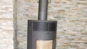A wood-burning stove with a chimney, set against a stone-patterned wall. - Langes Hus 2 - Ferienhäuser Langes Hus (Friedrichskoog)