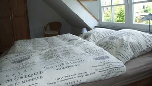 A bedroom with a sloped ceiling, a wooden wardrobe, a single chair, and a bed with a white duvet featuring printed text. - Langes Hus 2 - Ferienhäuser Langes Hus (Friedrichskoog)