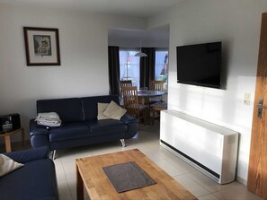 Un salón con un sofá azul, una mesa de centro de madera, un televisor montado en la pared y un radiador blanco.
