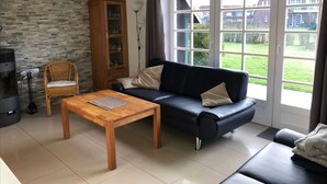 A living room with a sofa, a wooden table, a wicker chair, a cabinet, and a fireplace. - Langes Hus 2 - Ferienhäuser Langes Hus (Friedrichskoog)