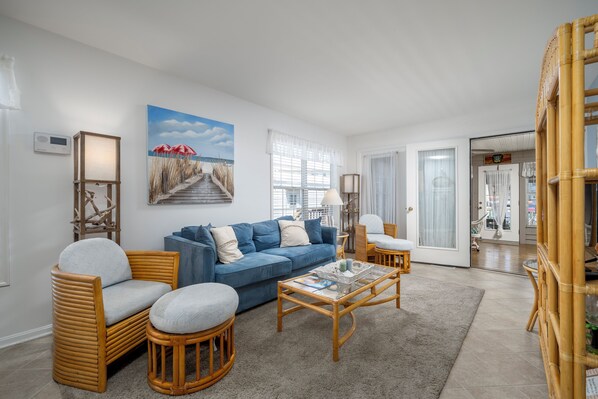 A living room with a blue sofa, wicker chairs, a coffee table, and a painting of a beach scene. - Outdoor Living At it's Finest With this Vacation Home! (Ocean City)