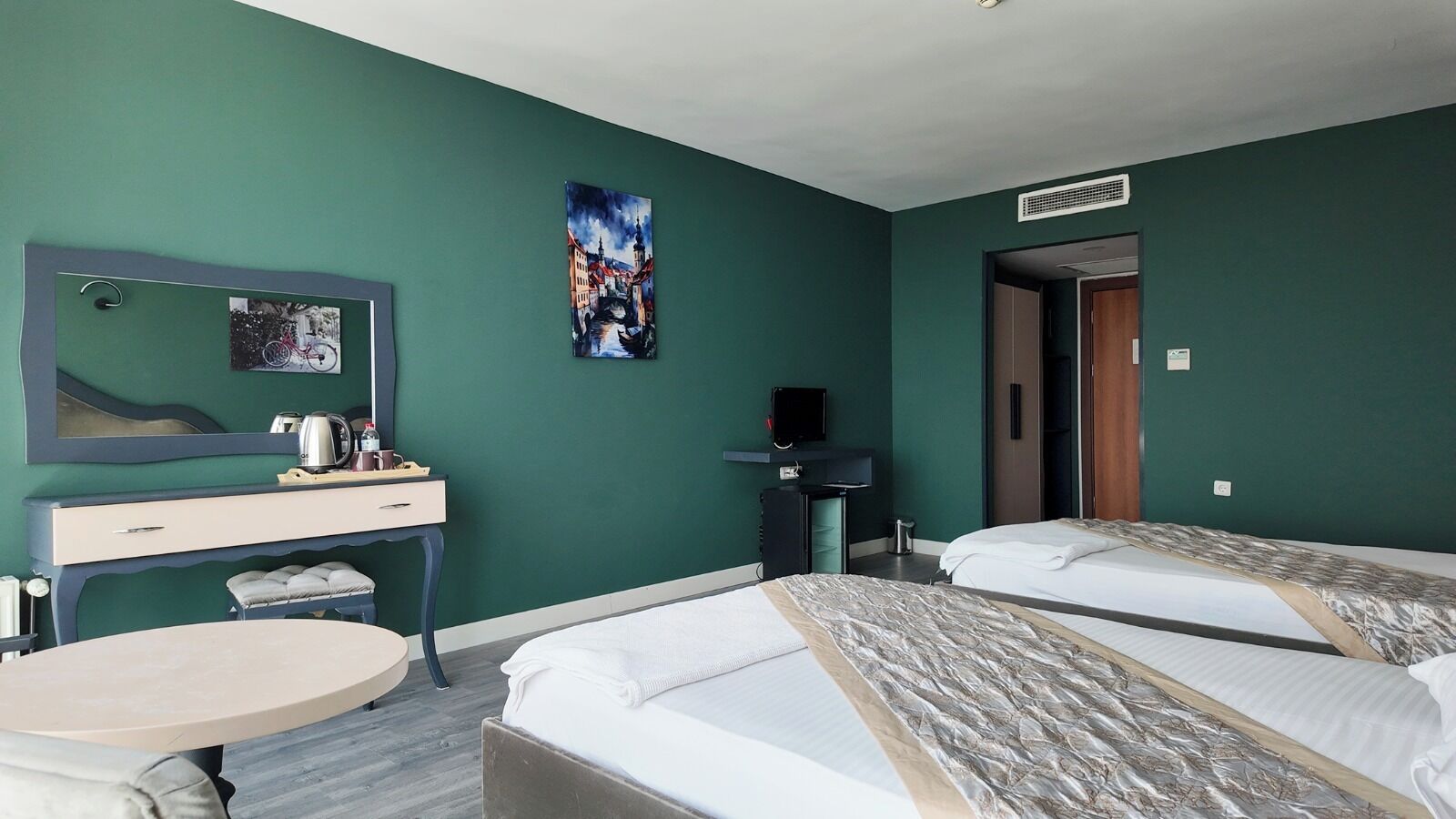 A hotel room with two beds, a desk with a chair, a small round table, a mirror, and a painting on the wall.