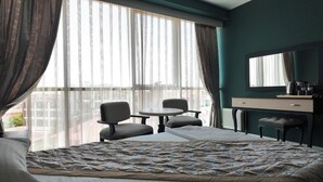 A hotel room with a large bed, two armchairs, a small table, a desk with a mirror, and a view of the city through large windows. - Grand Merpond Otel (Merzifon)