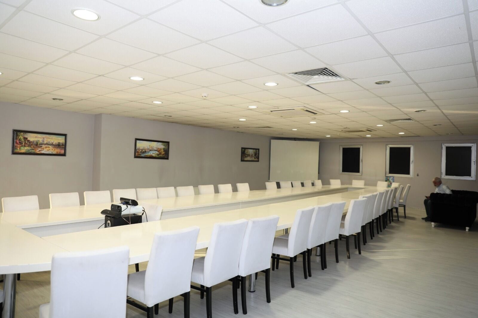 Un salón de conferencias con una larga mesa, sillas blancas, una pantalla de proyector y obras de arte en las paredes.