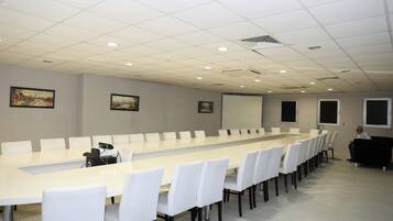 Un salón de conferencias con una larga mesa, sillas blancas, una pantalla de proyector y obras de arte en las paredes.
