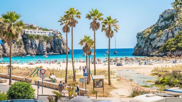 A beach with palm trees, a rocky cliff, and a building on the left. - ❤ BEACHFRONT & JACUZZI ❤ Casa 4Brothers (Cala en Porter)