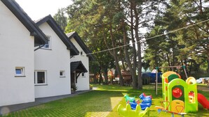 A children's play area with a white building, green and yellow play structures, and a grassy lawn.