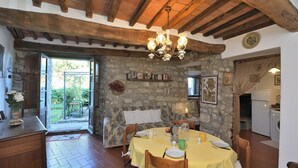 Una zona de menjador rústica amb una paret de pedra, bigues de fusta i un penjol. Hi ha una taula rodona preparada per dinar, una zona de cuina amb un frigorífic i una porta que fa a l'exterior.