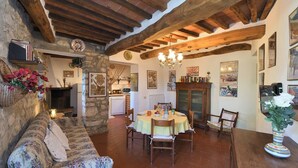 Un interior rústic amb bigues de fusta a la vista, una paret de pedra, una zona de menjador amb taula i cadires, i un espai d'estar acollidor amb un sofà.