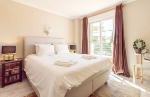 Uma cama bem arrumada com roupa de cama branca, uma mesa de cabeceira com abajur e uma janela com cortinas.