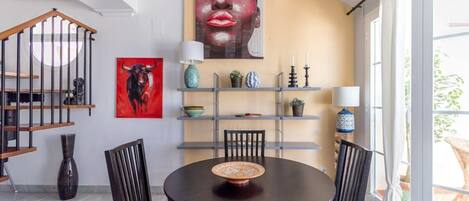A dining area with a round table, chairs, a wall-mounted shelf with decorative items, and a staircase.