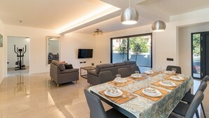 A modern dining area with a long table set for six, a sofa, and a TV in the background.